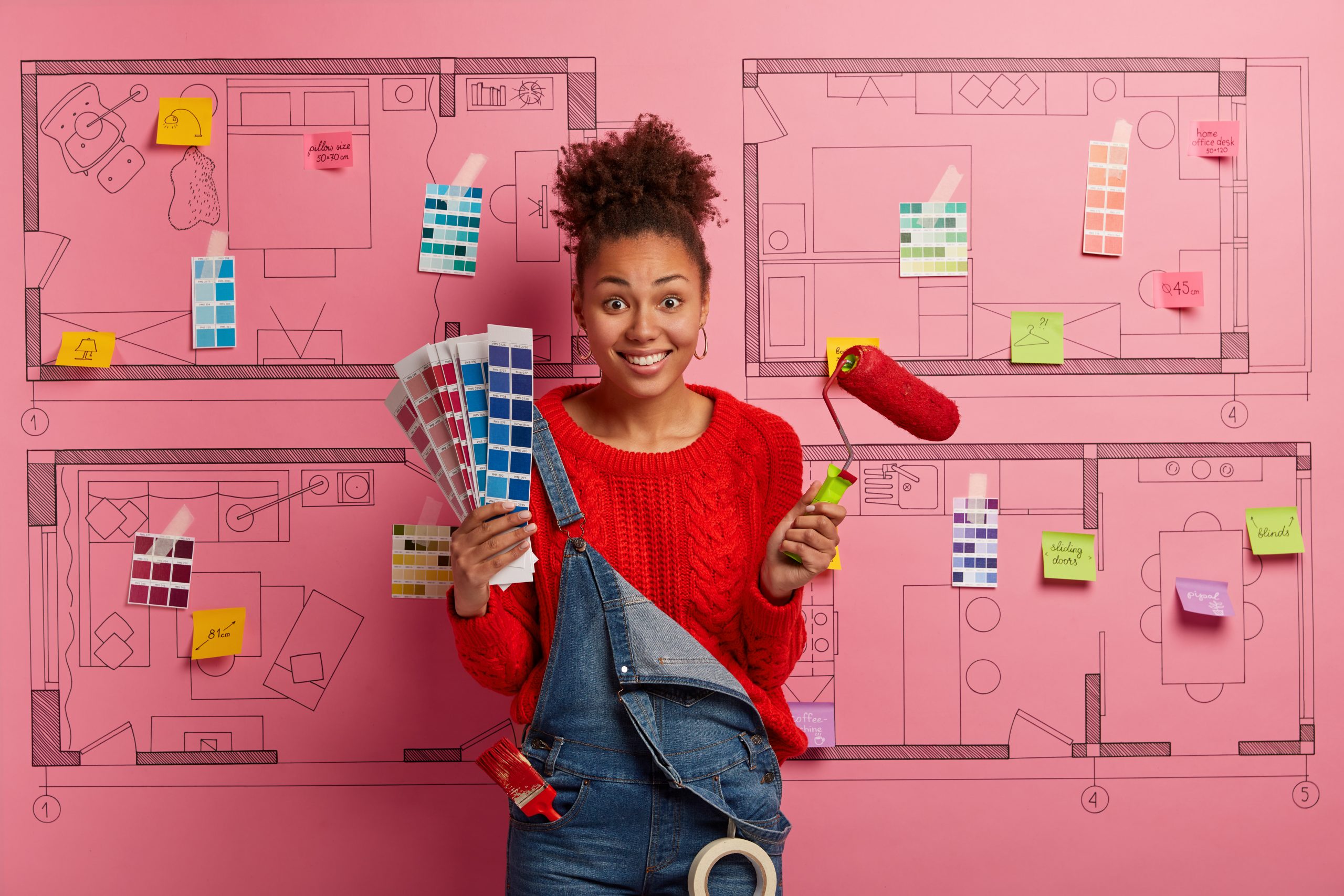 Glad skilled female designer stands with color samples and paint roller, ready for refurbishment in new house, wears red sweater and denim overalls, stands against apartment sketch design project